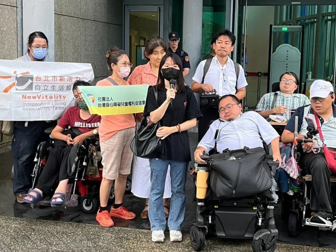 記者會近距離畫面,身障童盟專員沈昱綾手持麥克風發言,身旁站著多位與會者與一名坐電動輪椅的男士,背景展示「社團法人台灣身心障礙兒童權利促進會」布條。